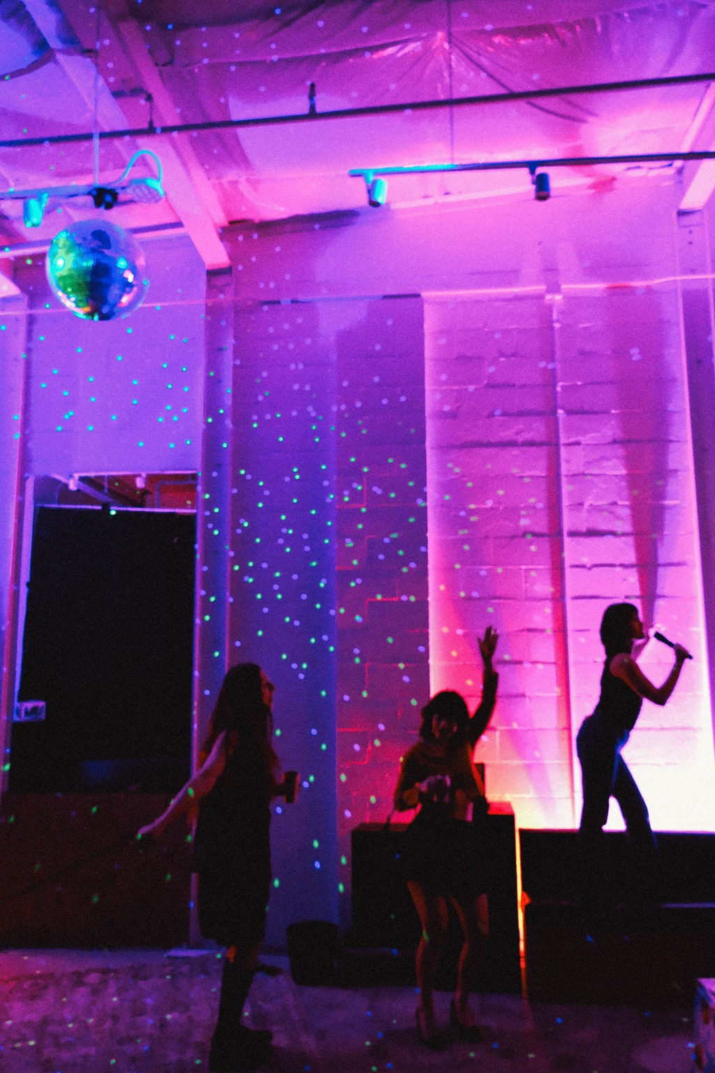 Silhouettes dancing under a disco ball and green laser dots in a purple-pink lit indoor loft party, with a singer on stage holding a microphone.