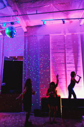 Silhouettes dancing under a disco ball and green laser dots in a purple-pink lit indoor loft party, with a singer on stage holding a microphone.