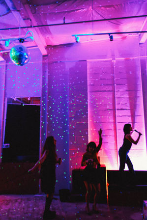 Silhouettes dancing under a disco ball and green laser dots in a purple-pink lit indoor loft party, with a singer on stage holding a microphone.