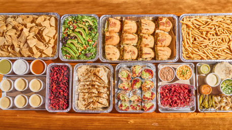 Vibrant catered Mexican-style spread on a wooden table — trays of tortilla chips, avocado salad, mini sandwiches, fries, quesadillas, tacos, rice, beans and assorted salsas and condiments.