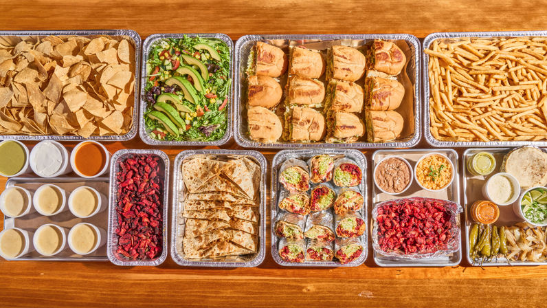 Vibrant catered Mexican-style spread on a wooden table — trays of tortilla chips, avocado salad, mini sandwiches, fries, quesadillas, tacos, rice, beans and assorted salsas and condiments.