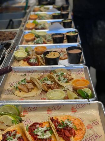 Row of metal trays on a restaurant counter with street-style Mexican tacos (al pastor and chicken) topped with onion and cilantro, served with lime wedges, salsa cups, and sides of rice and beans.