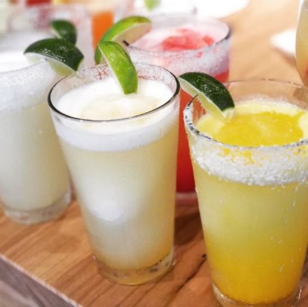 Colorful frozen margaritas on a wooden bar counter — yellow, white and pink icy cocktails with salted rims and lime wedges.