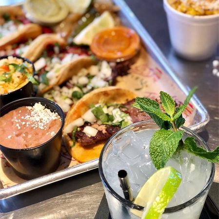 Close-up of Mexican street-style tacos on a metal tray with refried beans, dipping sauces and a zesty lime-and-mint cocktail