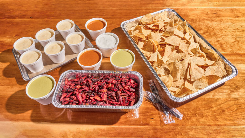 Takeout catering spread on a wooden table: large aluminum tray of tortilla chips, foil tray of chopped grilled steak, and several cups of queso, salsa roja, salsa verde and sour cream, ready for DIY nachos.