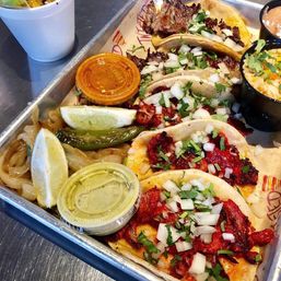 Vibrant tray of Mexican street tacos al pastor on corn tortillas, topped with chopped onion and cilantro, with lime wedges, grilled jalapeño, pickled onions and small containers of green and red salsa.