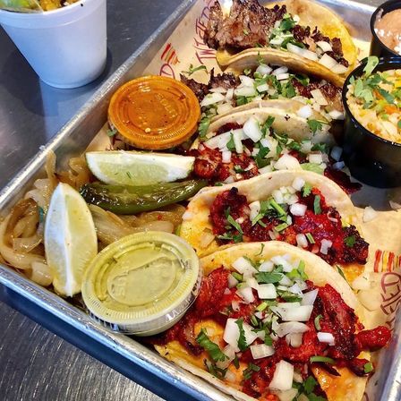 Vibrant tray of Mexican street tacos al pastor on corn tortillas, topped with chopped onion and cilantro, with lime wedges, grilled jalapeño, pickled onions and small containers of green and red salsa.