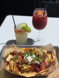 Sunlit tray of loaded Mexican-style nachos topped with shredded red meat, cilantro, queso fresco and a dollop of sour cream, paired with a chili-rimmed cucumber-lime margarita and a red fruit cocktail on a white patio table.