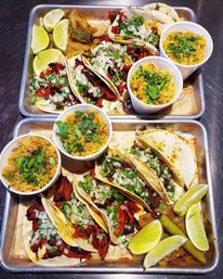 Two metal trays of Mexican street-style tacos al pastor on warm corn tortillas topped with diced onion and cilantro, served with lime wedges, pickled jalapeños and cups of cilantro-laced Mexican rice.