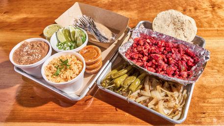 Street-style Mexican al pastor platter on a metal tray: foil of chopped red pork al pastor, grilled onions, roasted green chiles, corn tortillas, plus cups of Mexican rice, refried beans, lime wedges, chopped onion and cilantro, and small salsa containers on a wooden table.