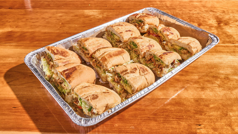 Catering tray of toasted pressed sandwiches cut into portions, filled with melted cheese, shredded lettuce and savory meat, on a rustic wooden table.
