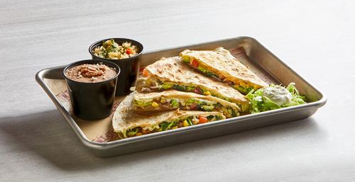 Grilled Mexican-style quesadilla wedges on a metal tray with melted cheese, corn, diced tomatoes and greens, served with cups of refried beans and seasoned rice, shredded lettuce and a dollop of sour cream.