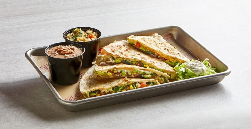 Grilled Mexican-style quesadilla wedges on a metal tray with melted cheese, corn, diced tomatoes and greens, served with cups of refried beans and seasoned rice, shredded lettuce and a dollop of sour cream.