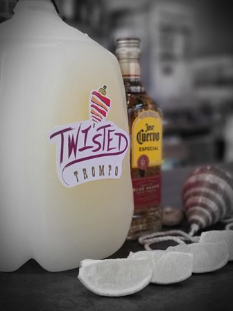 Plastic jug with a colorful "Twisted Trompo" sticker beside a tequila bottle, lime wedges and a striped spinning top on a bar counter