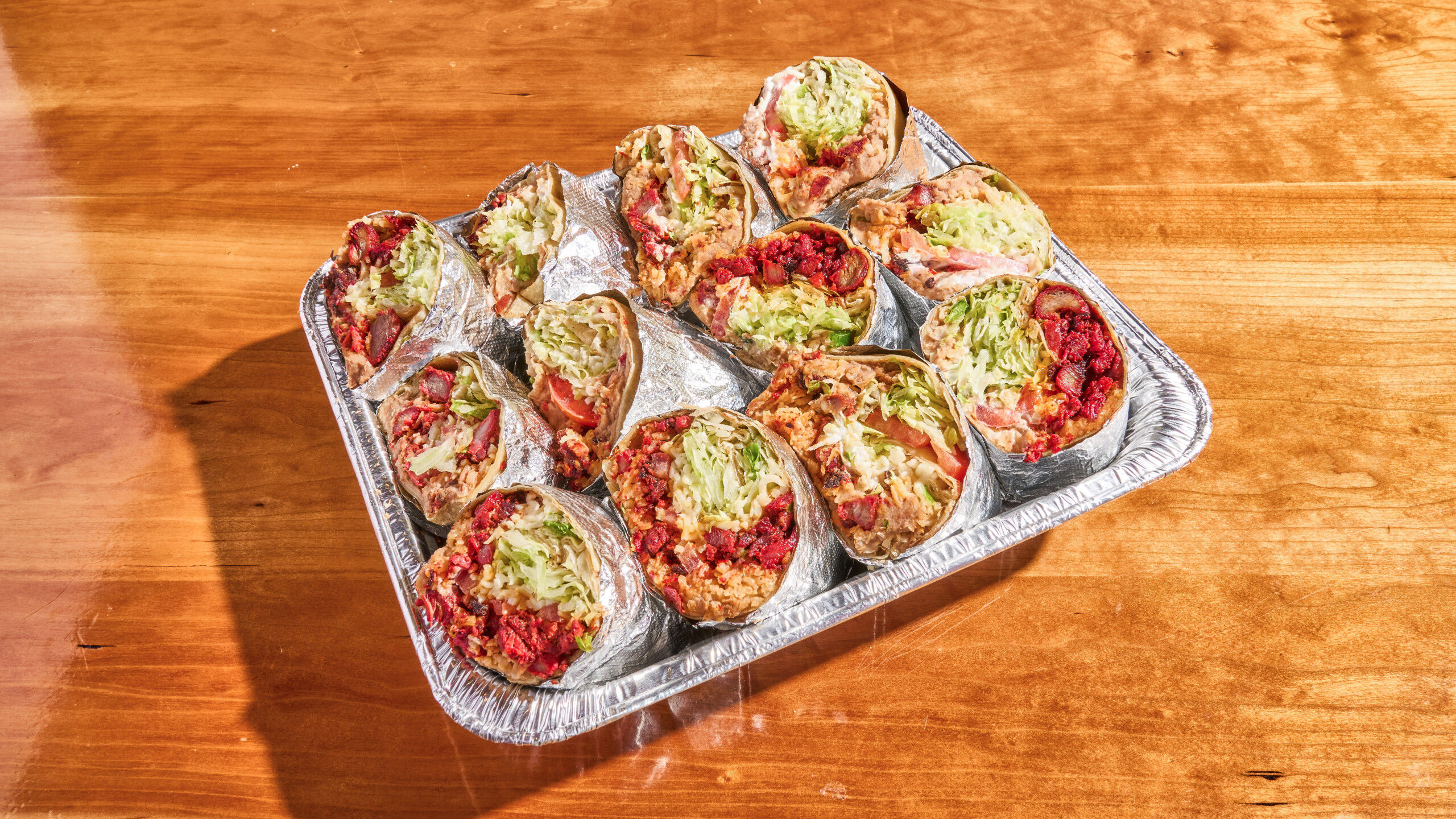 Aluminum takeout tray of halved foil-wrapped burritos bursting with rice, shredded lettuce, tomatoes and spicy red salsa on a wooden table
