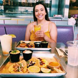 Smiling woman in a yellow sundress holding a citrus cocktail at a casual taqueria table with trays of street tacos, lime wedges, salsas, side dishes and drinks.