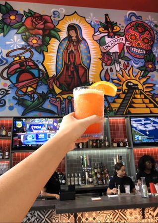 Hand holding an orange cocktail with an orange slice in a Mexican‑inspired bar, colorful mural of the Virgin of Guadalupe, sugar skull, roses and sun/pyramid above the bar with bartenders and TV screens.
