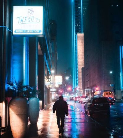 Lone pedestrian walking down a rainy, neon-lit downtown city street at night with blue and pink reflections on wet pavement, tall buildings and traffic in the background.
