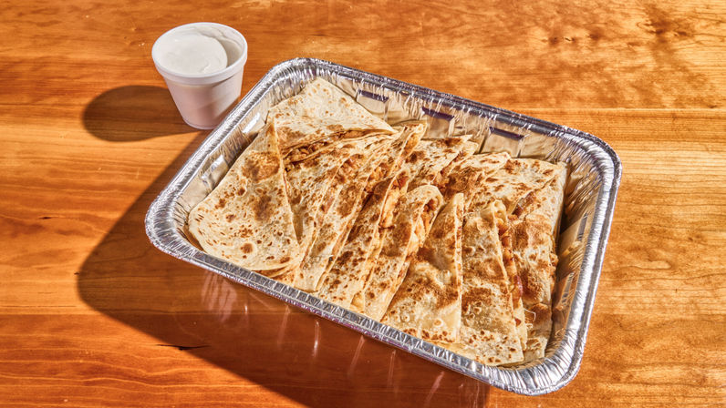 Golden, crispy quesadilla wedges in an aluminum takeout tray with melted cheese peeking out, served with a cup of sour cream on a wooden table.