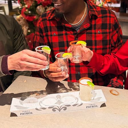 Three people clinking salt‑rimmed tequila shot glasses garnished with lime wedges over a tasting tray at a festive bar, one person wearing a red plaid shirt and smiling.