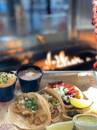 Three Mexican street tacos on a tray topped with cilantro and onions, served with rice, refried beans, lime wedges, pickled jalapeño and green salsa, blurred open‑flame grill in the background
