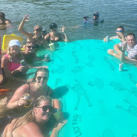 Group of adults relaxing around a large teal floating pad with frog prints in a calm lake, holding canned drinks and waving during a sunny summer lake party