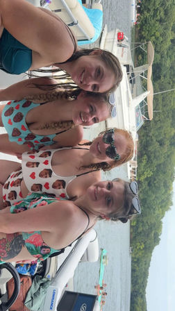 Four friends smiling on a pontoon boat during a summer lake day, wearing colorful swimsuits and sunglasses with wooded shoreline and other boats in the background.