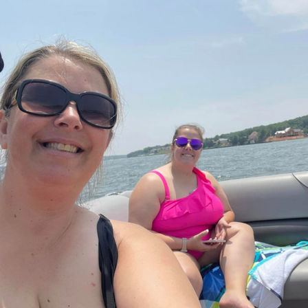 Two smiling women in swimsuits and sunglasses relaxing on a boat on a sunny lake with tree-lined shoreline and waterfront homes in the background