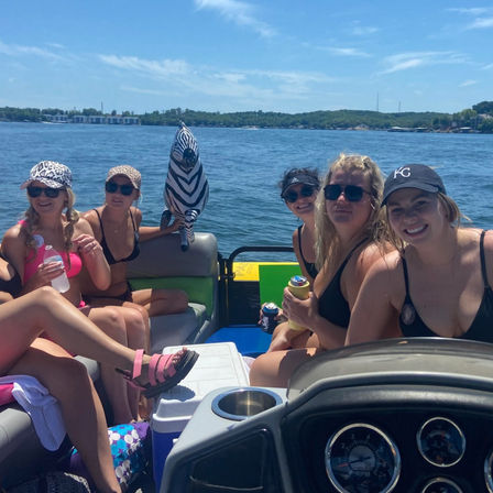 Group of friends on a pontoon boat enjoying a sunny lake day in swimsuits and sunglasses, holding drinks with a zebra-striped inflatable and shoreline in the background.