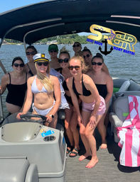 Cheerful group of friends on a pontoon boat at a sunny lake — women in swimsuits and sunglasses posing by the helm with a captain’s hat, shoreline and trees in the background for a summer boat rental outing.