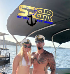 Two people in captain hats and sunglasses pose on a pontoon boat at a sunny lake dock, canopy displays a gold anchor boat-rental logo overhead.