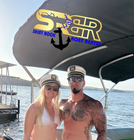 Two people in captain hats and sunglasses pose on a pontoon boat at a sunny lake dock, canopy displays a gold anchor boat-rental logo overhead.