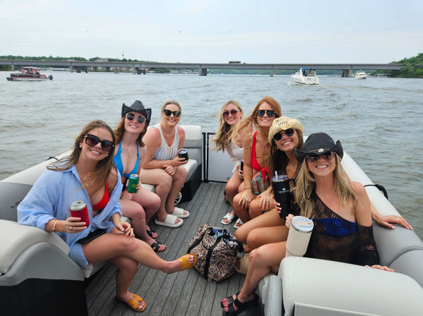 Eight women in swimsuits and sunglasses smiling and holding drinks on a pontoon boat, enjoying a summer river cruise with a bridge and other boats in the background.