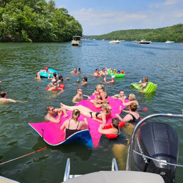 Group of people relaxing on a bright pink inflatable raft and assorted tubes in a lake with forested shoreline, boats and pontoons on a sunny summer day
