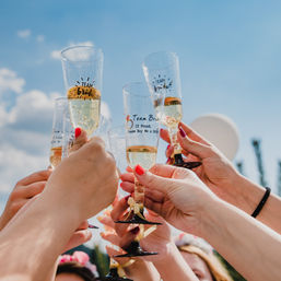 Hands with colorful nail polish raising 'Team Bride' champagne flutes in a sunny outdoor bachelorette party champagne toast against a bright blue sky