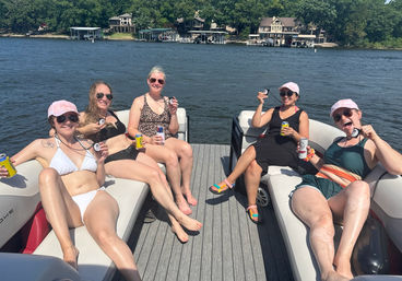 Group of five women relaxing on a pontoon boat on a sunny lake, wearing swimsuits and sunglasses and holding canned drinks, with tree-lined shoreline and waterfront homes in the background.