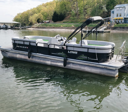 Black-and-gray pontoon boat with folded canopy moored on a calm lake, green water reflecting a wooded shoreline and lakeside docks and houses.