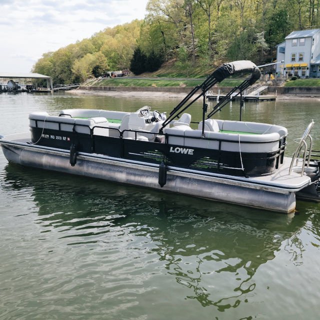 Black-and-gray pontoon boat with folded canopy moored on a calm lake, green water reflecting a wooded shoreline and lakeside docks and houses.