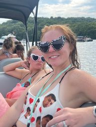 Two friends relaxing on a pontoon boat at a summer lake — one in rhinestone "bride-to-be" cat-eye sunglasses and printed swimsuit, the other in white heart-shaped shades, forested shoreline and boats behind them.