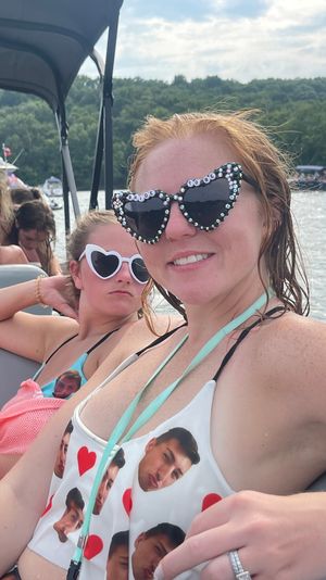 Two women relaxing on a boat at a forested lake on a sunny day, one wearing rhinestone 'Bride to Be' heart sunglasses and a novelty face-print swimsuit.