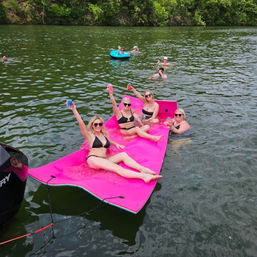 Group of women in black bikinis cheering with drinks while lounging on a bright pink floating mat tied to a boat on a green lake, with other swimmers and a blue tube near a tree-lined shore.