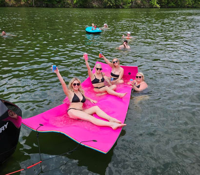 Group of women in black bikinis cheering with drinks while lounging on a bright pink floating mat tied to a boat on a green lake, with other swimmers and a blue tube near a tree-lined shore.