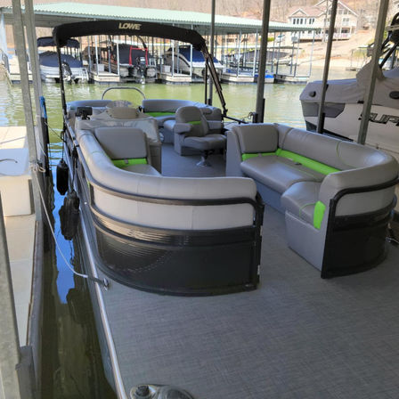 Pontoon boat docked in a covered marina slip on a calm lake, featuring spacious gray and lime-green cushioned seating, swivel captain's chair, open deck and bimini top.