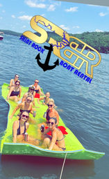 Group of people relaxing on a bright green floating mat in a sunny lake, holding canned drinks; tree-lined shore and blue sky in background with an overlaid anchor and boat rental logo graphic.