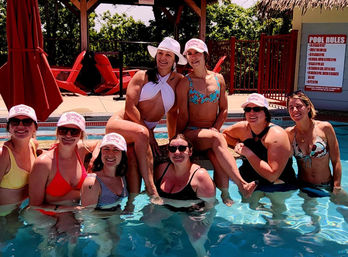Eight women in colorful swimsuits and matching pink caps smiling and posing together at an outdoor pool for a bachelorette-style summer pool party, red lounge chairs and a pool rules sign in the background.