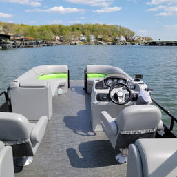 Pontoon boat interior with gray and lime-green seating and helm, cruising on a calm lake toward a tree-lined shore with waterfront homes, docks, and a sunny blue sky.
