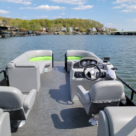 Pontoon boat interior with gray and lime-green seating and helm, cruising on a calm lake toward a tree-lined shore with waterfront homes, docks, and a sunny blue sky.
