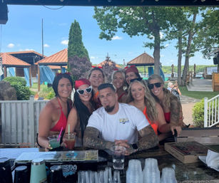 Cheerful group of friends in swimsuits and summer outfits gathered around a tattooed man at a lakeside outdoor bar patio, with cabanas, trees, and a dock visible under a sunny blue sky — summer lakefront hangout.