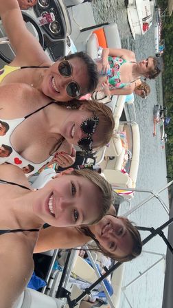 Four friends smiling in swimsuits and sunglasses on a pontoon boat on a sunny lake, surrounded by other boats and a tree-lined shoreline.