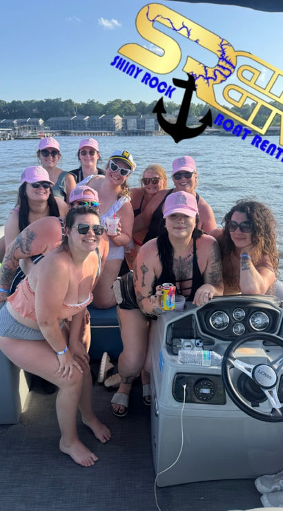 Group of women in swimsuits and matching pink caps enjoying a sunny pontoon boat party on a lake, holding drinks near the boat helm with lake houses and a tree-lined shore in the background.